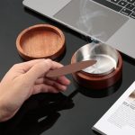 Hand met sigaret bij een houten asbak met metalen inzet, op een bureau.