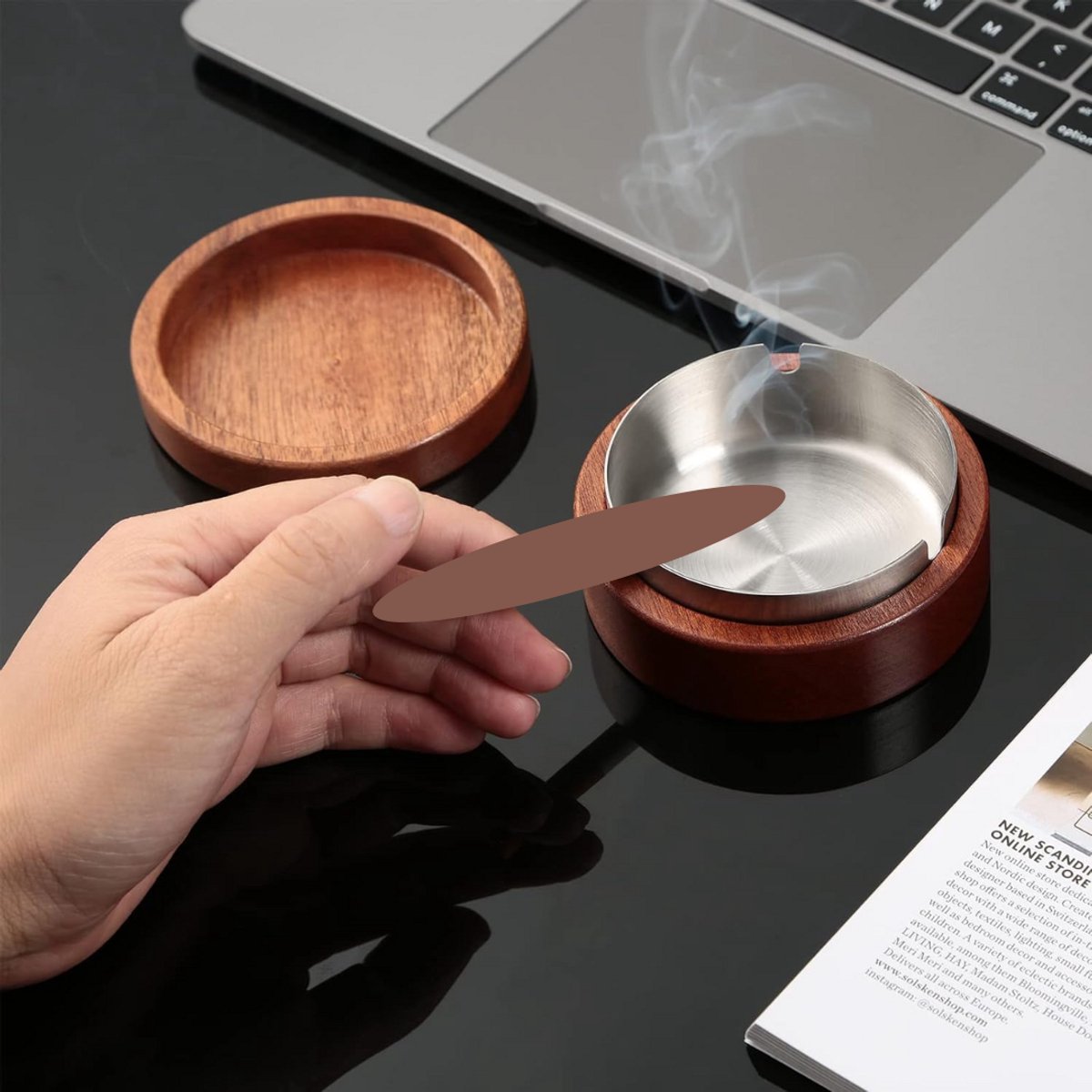 Hand met sigaret bij een houten asbak met metalen inzet, op een bureau.
