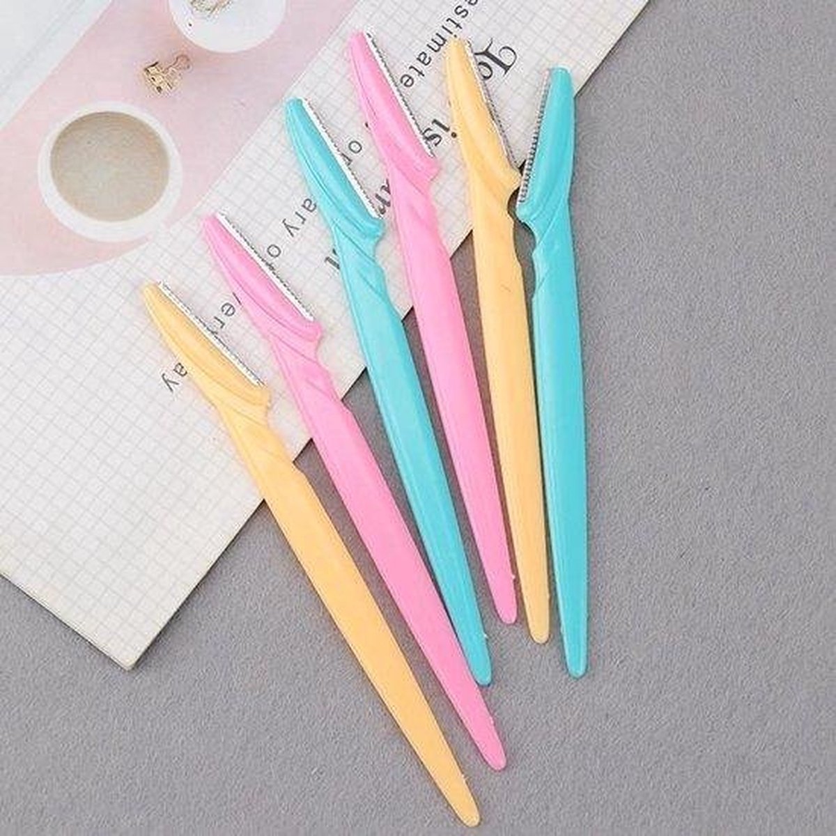 Zes gezichts-scheermesjes in pastelkleuren roze, turquoise en geel, op papier.